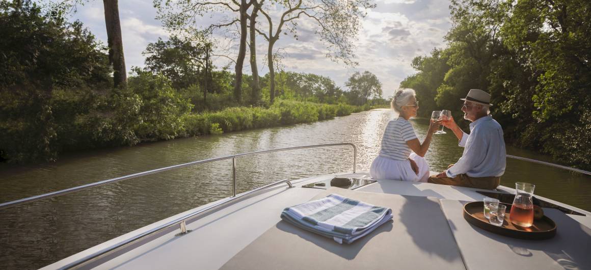 Un couple profite d'un moment romantique au coucher du soleil, en buvant un verre à l'avant de leur bateau, sur un canal calme.